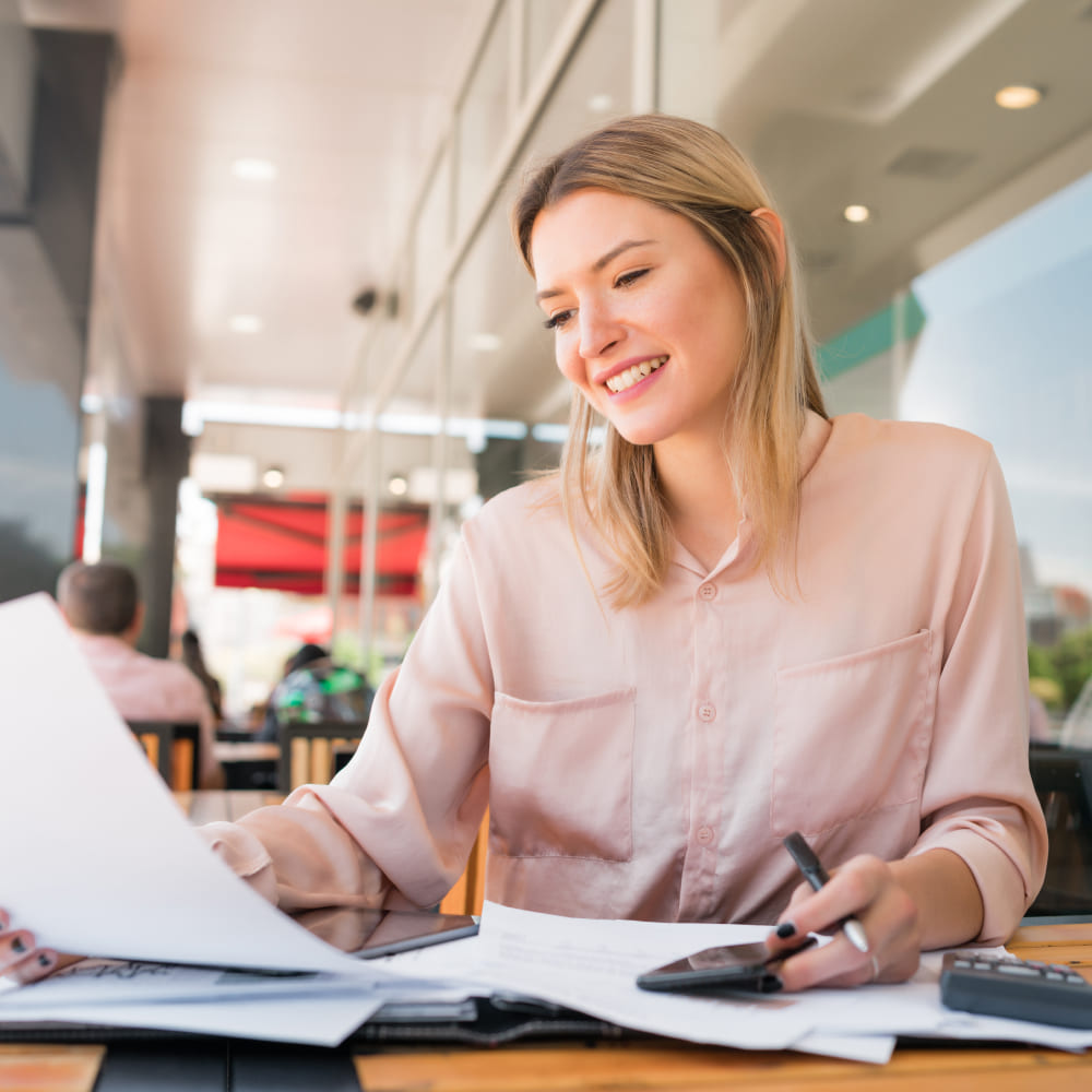 Een jonge vrouw werkt met documenten en een rekenmachine in een café, glimlachend en geconcentreerd.